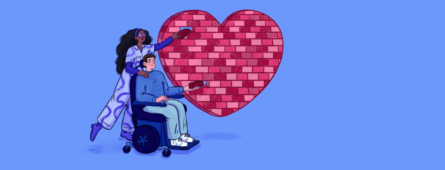 A woman stands behind a man in a motorized wheelchair, they are building a heart made of blocks together
