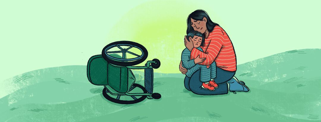 A woman sits on the ground and holds a child with a scraped knee in her lap next to a tipped over wheelchair